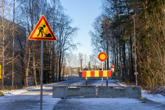 A road is blocked by concrete barriers with traffic signs, including a roadwork warning sign, indicating construction and restricted access to the area