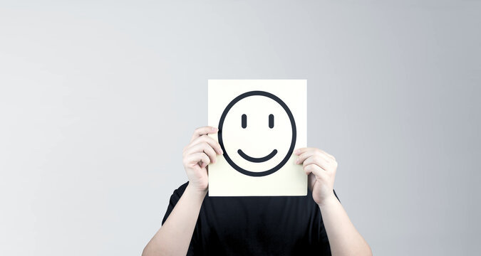 Anonymous man holds paper with smiley face emoji covering his face, standing on white background. Concept of mental health assessment, or rating the level of happiness.