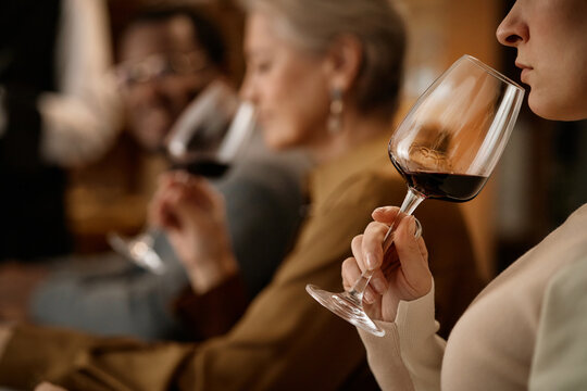 Middle aged Caucasian woman holding wine glass near face, senior Caucasian woman and middle aged Black man sitting in background, all smelling and tasting red wine during testing
