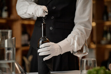 Caucasian young adult man wearing formal attire opening wine bottle with corkscrew, hands covered in gloves, preparing for wine tasting event, empty wine glass nearby