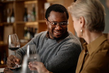 Middle aged Black man smiling and holding wine glass while sitting across from middle aged Caucasian woman tasting wine in modern tasting room, both engaging in wine evaluation