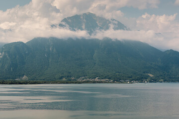 Peaceful view of Lake Geneva with towering green mountains, misty clouds, and a quiet lakeside town nestled at the base of the slopes, Swiss Alps, Switzerland.
