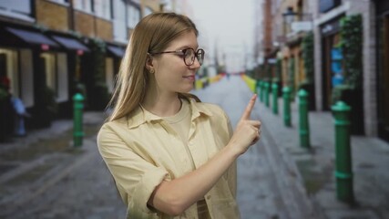 Blonde woman wearing glasses and beige shirt pointing finger on sunlit city street with cobblestone pavement and green bollards; confidence curiosity guidance direction.