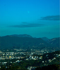 A moody, dramatic view of the Sauk suburb of Tirana and the surrounding hills, featuring the moon visible in a deep turquoise sky above the mountains.