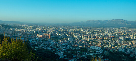 A sweeping panoramic view of the vast Tirana cityscape extending toward the horizon, backed by a series of low mountains under a hazy, bright blue sky.