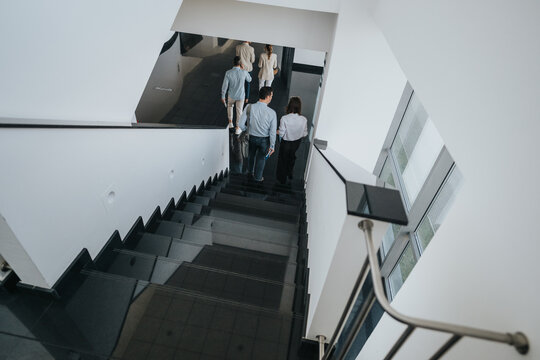 A group of people walk down sleek stairs in a clean, white-walled interior. Natural light from windows highlights glossy steps and glass railings, creating a professional yet calm office atmosphere. - Powered by Adobe