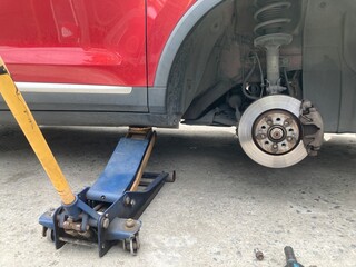 Low-angle view of a red car lifted by a hydraulic jack on a concrete floor. The wheel is removed, showing the brake disc and caliper assembly during a DIY repair or maintenance process.