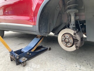 Low-angle view of a red car lifted by a hydraulic jack on a concrete floor. The wheel is removed, showing the brake disc and caliper assembly during a DIY repair or maintenance process.