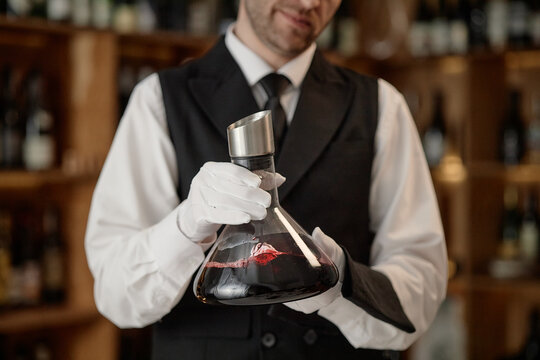 Caucasian middle aged man wearing formal vest and gloves holding glass wine decanter, carefully inspecting red wine in upscale wine cellar, performing professional wine testing