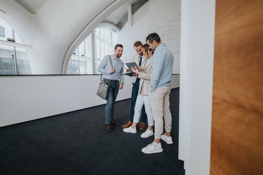 A diverse group of coworkers and associates gathers in a bright, contemporary hallway to review a tablet, share ideas, and plan next steps.
