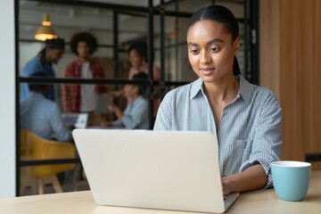 Woman working on laptop in modern office with colleagues collaborating in the background behind her