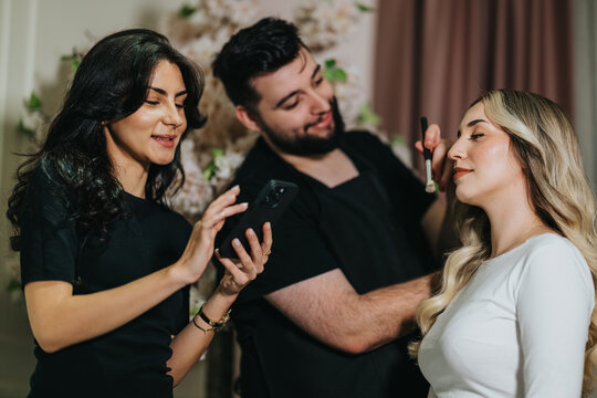 Three friends share a relaxed moment in a hair studio as one applies makeup and others view on a phone, creating a warm, collaborative beauty scene.