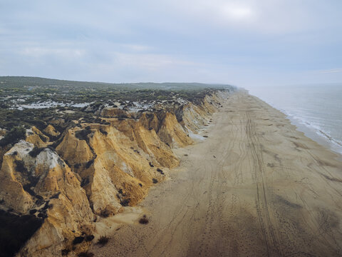 Aerial view of a dramatic coastline where golden cliffs meet the sandy shore under a serene sky, Playa Torre del Loro, Mazagon, Andalusia, Spain.