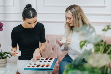 Two friends share a relaxed moment, sipping tea and exploring a chocolate tasting board on a pink...