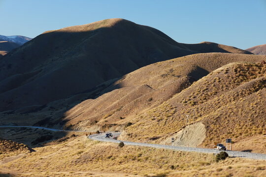 Mackenzie Basin, New Zealand - May 23, 2025: Dry, tussock-covered rolling hills and mountains on the road to Mount Cook. Sunlight and shadow emphasize the dramatic, vast landscape, with a winding road