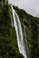 Fototapeta premium Milford Sound, New Zealand - May 20, 2025: A massive waterfall cascades down the sheer, moss-covered cliff face of the fjord. The scene is shrouded in mist and fog, typical of the Fiordland landscape.