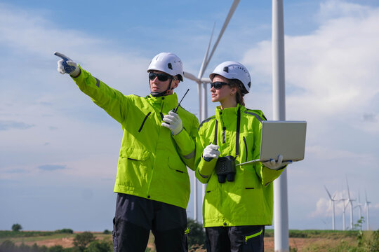 Engineers inspecting wind turbines with laptop in bright safety jackets during sunny day in a renewable energy farm