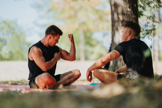 Two friends share a casual outdoor workout in the park. One flexes his bicep while the other chats nearby, capturing fitness, camaraderie, and motivation in natural light.
