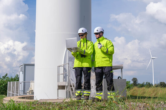 Engineers inspect wind turbine site while using laptop in bright safety gear on a clear day