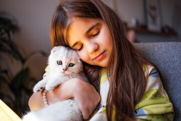 Young girl with long hair gently cuddling a fluffy white kitten, showcasing a tender moment of...