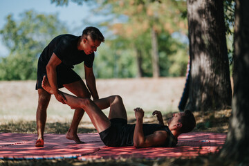 Two athletic men perform a partner leg stretch on a red mat in a sunny park, one standing and assisting the other lying down. Focus on fitness, balance, and teamwork.
