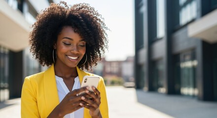Happy African American woman using smartphone in city street. Smiling professional lady in yellow blazer texting on mobile phone outdoors. Communication concept