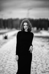 Elegant woman in a long black dress stands confidently on a snowy pathway, surrounded by winter...