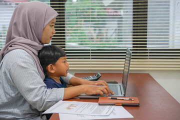 Side view of mother wearing hijab working on laptop with her child on her lap, remote work concept with notebook, documents, pen, and calculator on the table