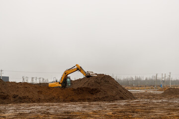 A yellow crawler excavator works with a large pile of soil on a construction site in cloudy weather
