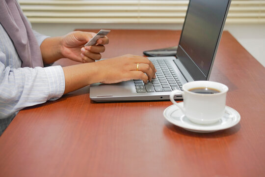Woman using a laptop and holding a credit card for online payment on a wooden desk with coffee in a home workspace