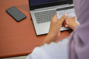 Top view of a woman holding a credit card, blurred in the foreground, with focus on laptop keyboard and smartphone on a brown desk, copy space included for design