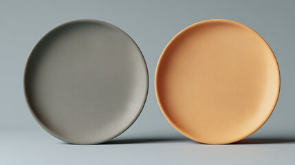 Two minimalist plates. One is a shade of gray, the other is light orange, set against a simple backdrop.