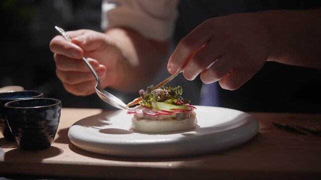 A chef is preparing a dish with a spoon and a knife. The dish is on a white plate and has a garnish of vegetables. The chef is focused on the task at hand, and the scene conveys a sense of precision