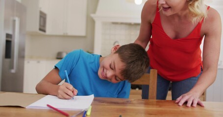 Drawing blue-shirt boy leaning over notebook at kitchen table, woman red-top watching with blue pen