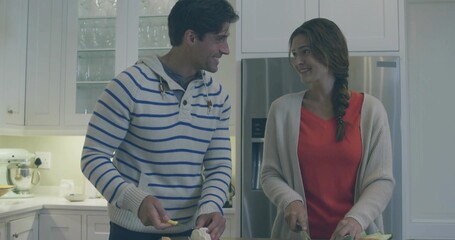 Chopping couple wearing red top and striped sweater locking eyes at kitchen island, using knives