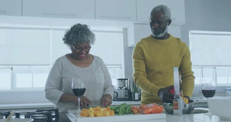 Chopping woman in gray top at kitchen island, man in mustard sweater rinsing produce, wine glasses