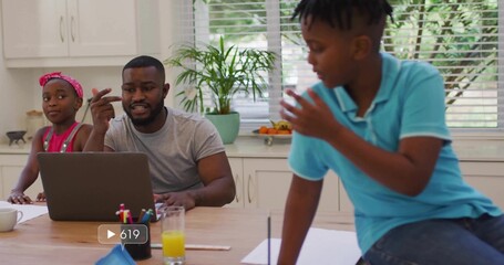 Working father gray tee typing on laptop kitchen table, daughter pink headband son light blue polo