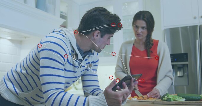 Leaning man in striped sweater checking phone in kitchen, woman chopping vegetables with knife - Powered by Adobe