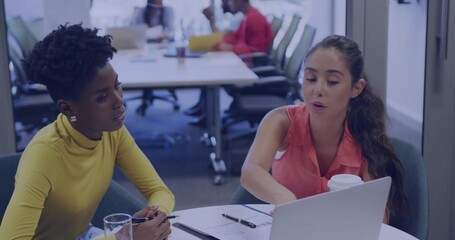 Discussing women wearing mustard top and coral blouse, reaching laptop and holding coffee in office
