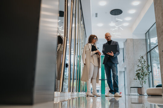 Two professionals review work details in a bright, contemporary office lobby. A man holds a tablet while a woman watches, suggesting collaboration, planning, and a productive business meeting. - Powered by Adobe