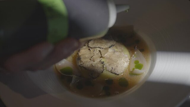 A person is pouring a white soup into a bowl. The soup is garnished with green peas and has a garnish of black truffle