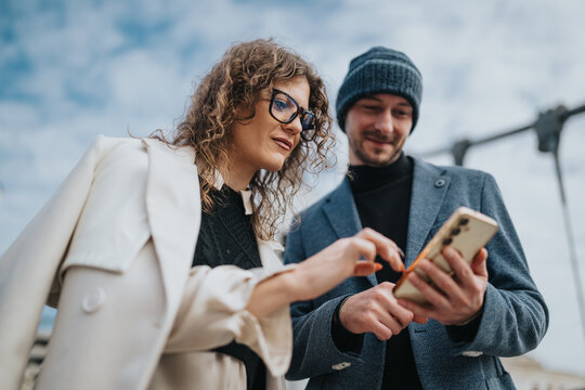 Two colleagues collaborate outdoors, examining a smartphone together. They wear smart jackets, showing a relaxed, professional vibe as they discuss ideas against a bright blue sky. - Powered by Adobe