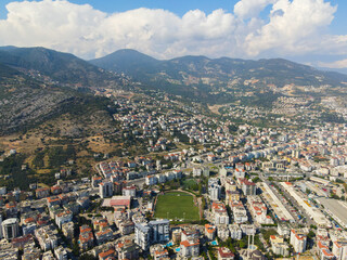 Wide aerial view showing the dense cityscape of Alanya, a football field, and the extensive residential development creeping up the steep mountain slopes
