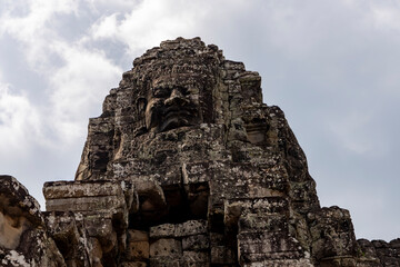 Close-up of weathered stone face carved into Bayon Temple tower. Cloudy sky in background. Mood: mysterious, solemn. Use: travel photography, cultural documentation, architectural study