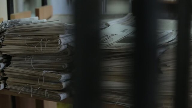 Piles of old archived newspapers stacked on shelves in a library storage room. The bundles are tied with string and seen through a railing during a pan shot.


