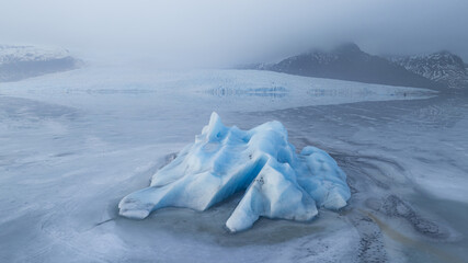 Aerial view of a striking blue iceberg adrift in the glacial lagoon, a serene dance of ice and water under a misty sky, Jokulsarlon, Sveitarfelagid Hornafjordur, Iceland.