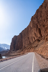 A wide shot of an asphalt road winding along the base of a towering, textured red-orange rock cliff face with a river and mountains visible in the distance