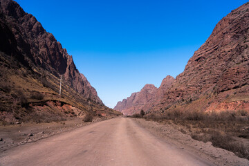 A straight dirt road leading deep into a narrow gorge flanked by towering, steep, red rock cliffs under a clear blue sky