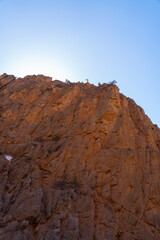 A dramatic vertical view of a towering, textured orange-brown rock cliff wall against a clear, bright blue sky in a canyon or mountain pass