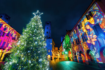An illuminated Christmas tree in the piazza, flanked by buildings adorned with native scenes light projections, during the festive season in Alba, Italy.